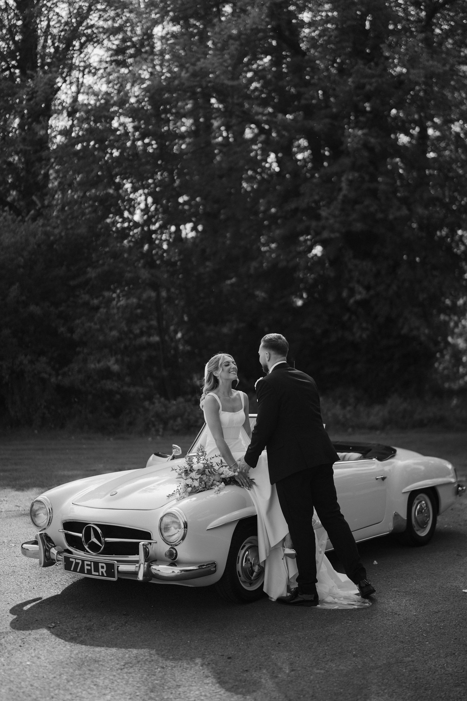 groom leaning in for a kiss with the bride as she sits on car bonnet at ivy lodge in cirencester