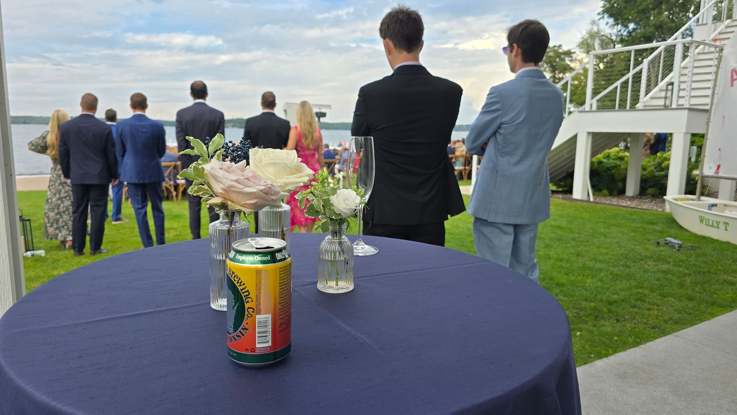 Guests gather on a green lawn overlooking water during an outdoor event with drinks on a navy blue cocktail table.