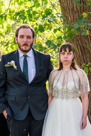 A couple in formal wedding attire poses together in a garden setting with green trees and foliage in the background.