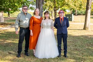 Wedding party members stand together outdoors in formal attire on a sunny autumn day in a park setting.