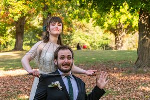 A couple in formal attire playfully poses together in a park setting with autumn leaves scattered on the ground.