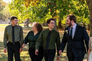 A wedding party in dark suits stands outdoors among autumn trees during a fall celebration.