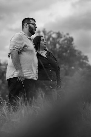 A romantic couple embraces in a moody black and white portrait amid tall grass and cloudy skies.