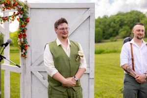 Two men in formal attire stand outdoors at a wedding ceremony, one wearing a green vest with a boutonniere.