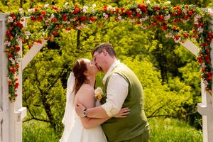 Bride and groom share a kiss under a floral arch adorned with red roses at an outdoor wedding ceremony.