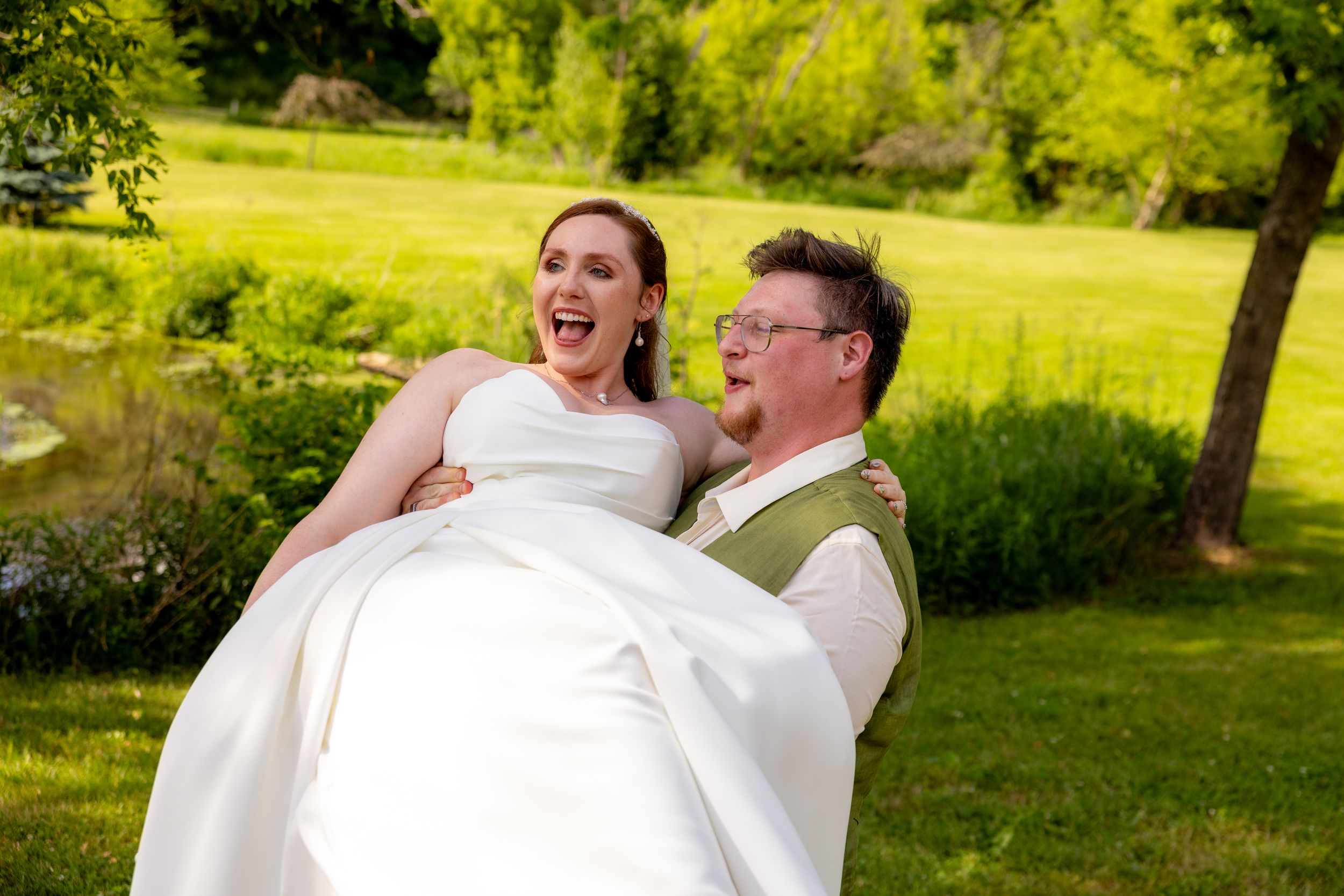 Joyful groom carrying laughing bride in white wedding dress outdoors on lush green lawn.