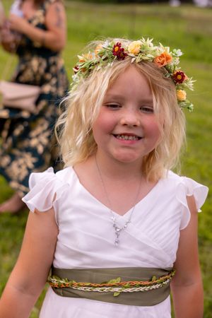 Smiling young flower girl in white dress with floral crown and olive sash at outdoor wedding ceremony.