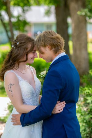Bride and groom in blue suit share a romantic forehead touch outdoors, surrounded by lush green trees.