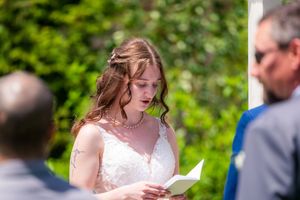 Bride in white lace dress reads vows from paper during outdoor wedding ceremony on sunny day.