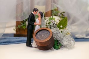 Wedding cake topper figurines of bride and groom beside engraved wooden ring box with flowers.