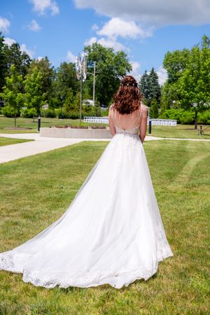 Bride in white lace wedding gown with long train standing in green park, viewed from behind on sunny day.