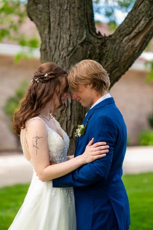 A bride and groom share an intimate forehead touch moment outdoors near a large tree, wearing wedding attire.