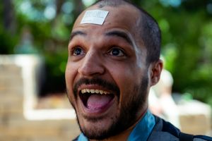Smiling man with a sticky note on his forehead, laughing outdoors in a lush green setting.
