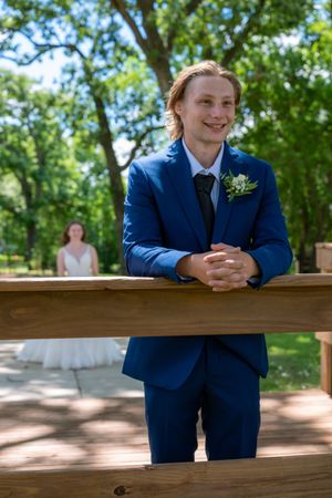 Smiling groom in blue suit with boutonniere leans on wooden fence, bride in white dress visible in background outdoors.