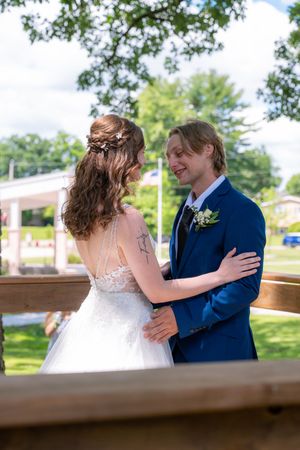 A smiling groom in a blue suit embraces his bride in a white lace dress outdoors on their wedding day.