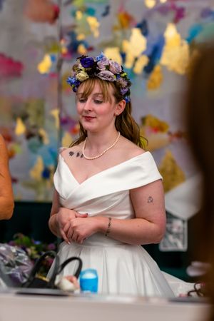 Bride in white off-shoulder gown and floral crown with purple flowers stands elegantly at a colorful art-filled venue.