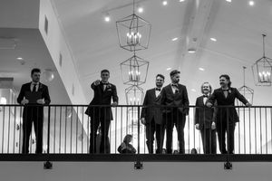 Groomsmen in tuxedos posing on a balcony with elegant chandeliers at a wedding venue.