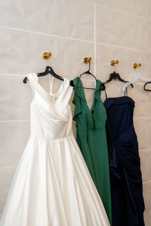 White wedding gown and green and navy bridesmaid dresses hanging on gold hooks against a white tiled wall.