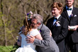 Father embracing bride in white dress and floral crown at outdoor wedding ceremony with groomsmen in black suits.