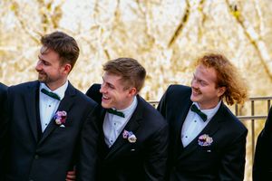 Three groomsmen in black tuxedos with floral boutonnieres laughing together outdoors at a wedding.