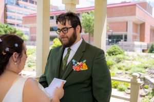 Groom in green suit with colorful pocket square listens as bride reads wedding vows at outdoor ceremony.