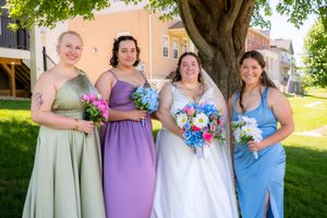 Bride in white gown with three bridesmaids in green, purple, and blue dresses holding bouquets outdoors.