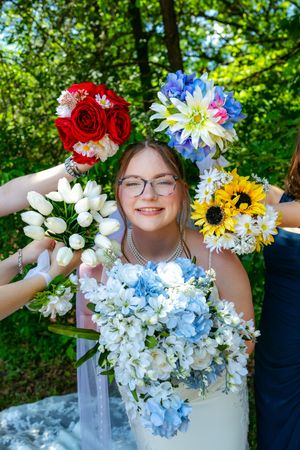 Smiling bride in white dress holds blue bouquet surrounded by colorful bridesmaid bouquets outdoors.
