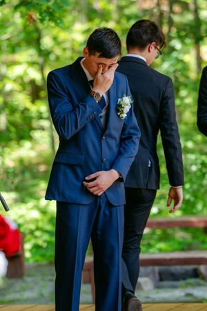 Emotional groom in blue suit with boutonniere wiping tears at outdoor wedding ceremony among green trees.