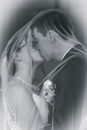 Black and white photo of a bride and groom sharing a romantic kiss under a flowing wedding veil.