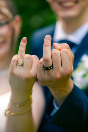 Newlyweds showing off wedding rings on middle fingers, bride in gold band and groom in black band, smiling outdoors.