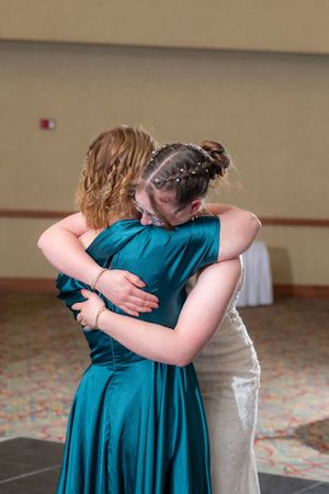 Bride in white gown and bridesmaid in teal dress share an emotional hug on the wedding reception dance floor.