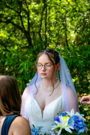 Bride in white lace gown, pearl necklace, and veil holds blue bouquet outdoors with lush green trees behind her.