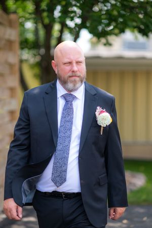 Bald bearded groom in dark suit with floral boutonniere and blue paisley tie walking outdoors.