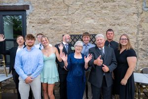 A joyful family group poses and waves outside a stone building, dressed in formal attire for a special occasion.