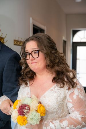 Emotional bride in white lace gown and glasses holding colorful bouquet of roses during wedding ceremony.