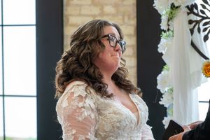 Bride in lace wedding dress and glasses standing at altar decorated with white flowers.