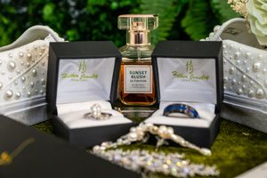 Elegant bridal flat lay with Sunset Blush perfume, diamond rings, blue band, pearl shoes, and sparkling jewelry on green backdrop.