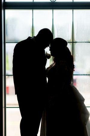 Romantic silhouette of a couple standing face to face against a large window, suggesting a wedding moment.