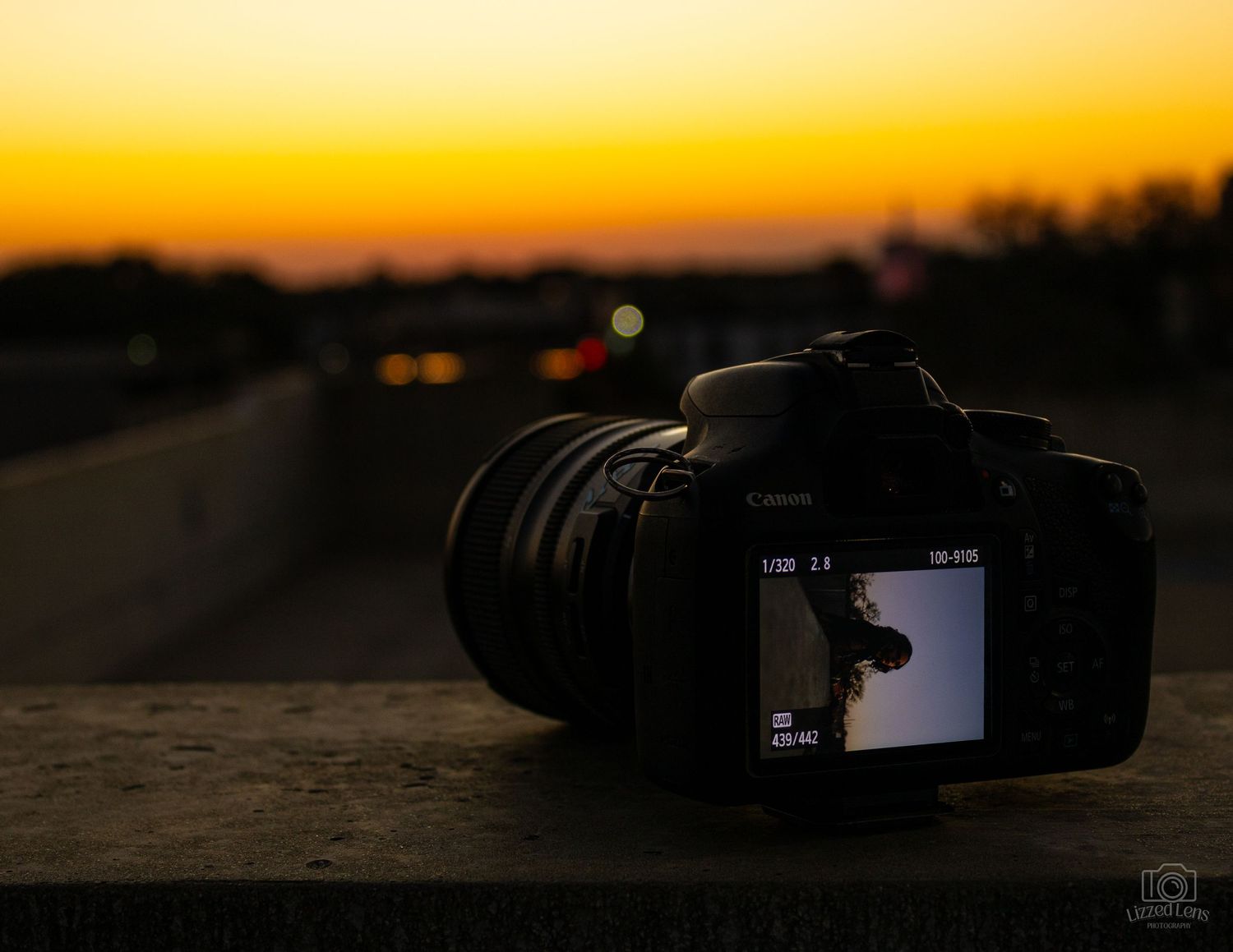Canon DSLR camera on a ledge at sunset with golden sky and bokeh city lights in the background.