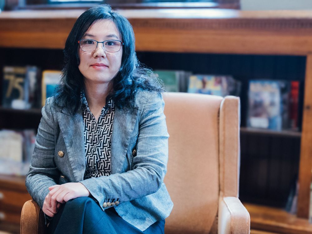 Someone wearing glasses and a gray blazer poses in a library setting.
