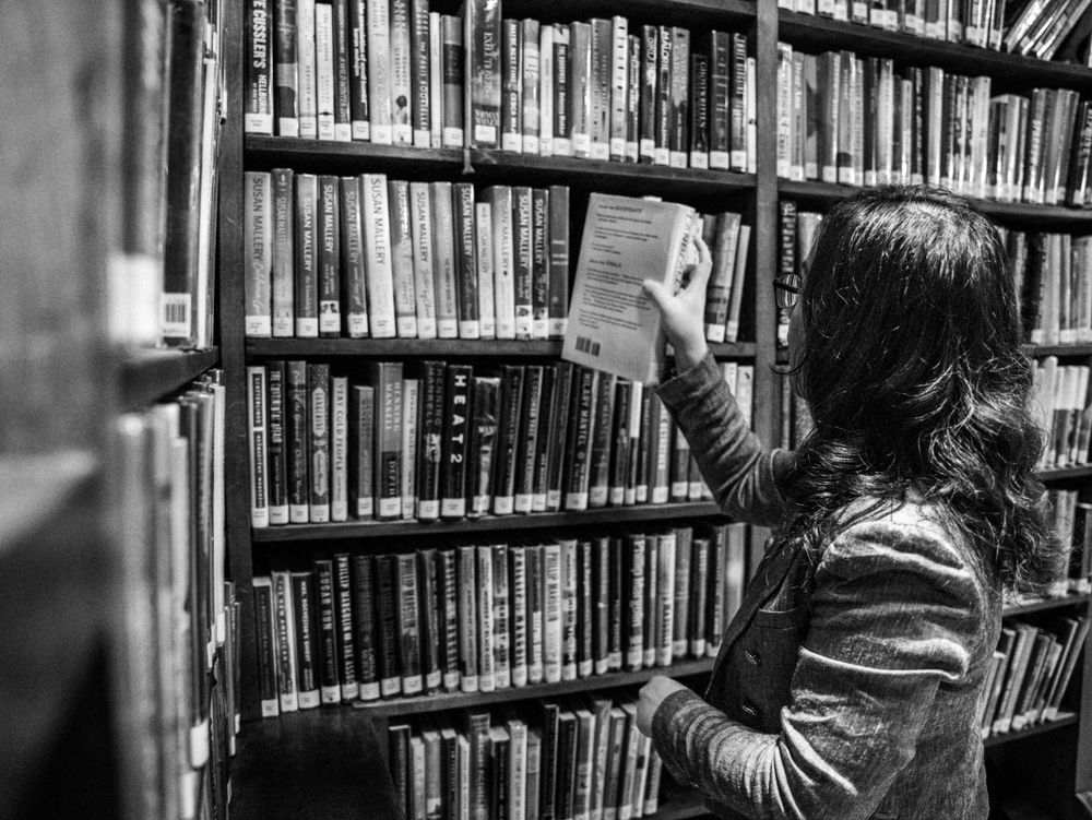 Someone reaches for a book while standing among tall library shelves in black and white.