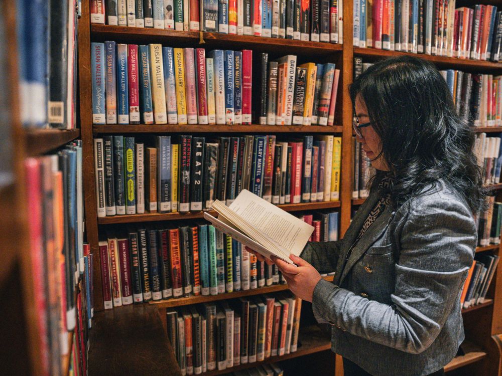 A reader in a grey jacket browses colorful library books on wooden shelving.