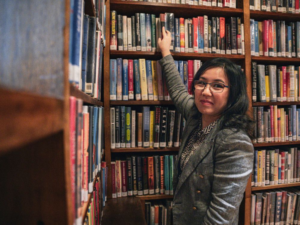A person reaches for books while standing among tall library shelves and wearing a grey blazer.