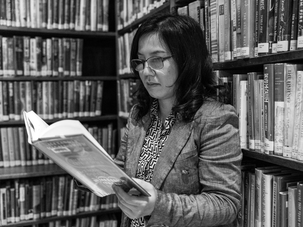 A reader in a grey blazer studies a book among library shelves in black and white.