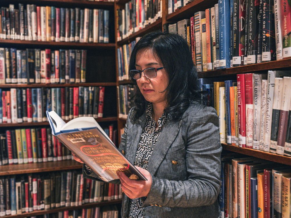 A person in professional attire reads intently while standing in a library's book stacks.