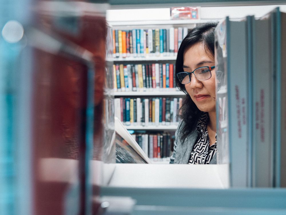 A reader peers through library shelves while holding a book in a creative composition.
