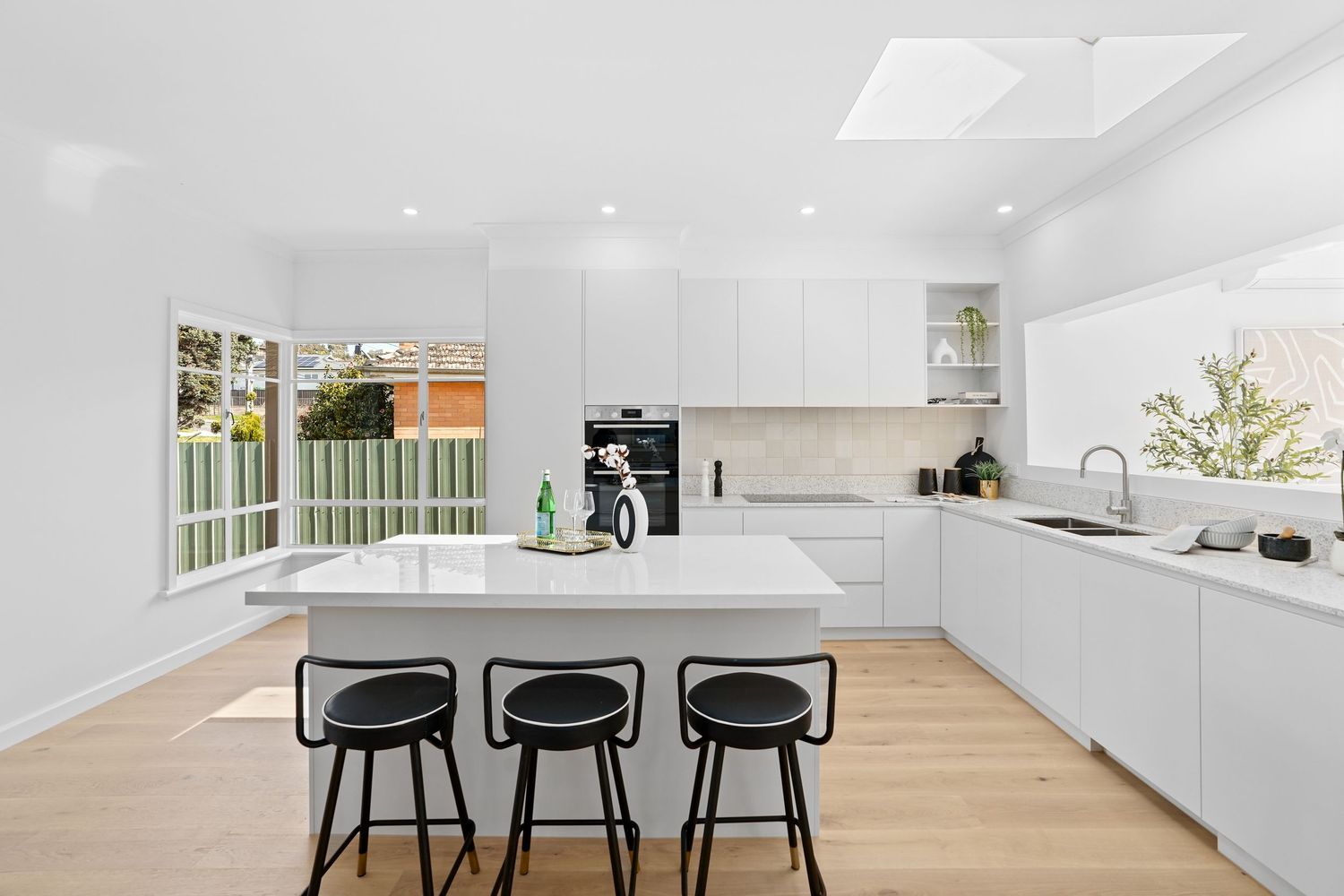 Modern white kitchen interior with wooden floor and black bar stools at a central island.