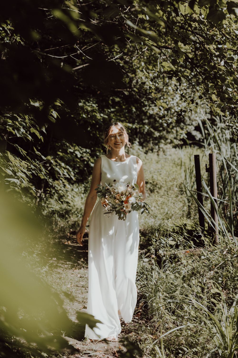 A bride in a flowing white dress walks through a sunlit garden path holding a bouquet.