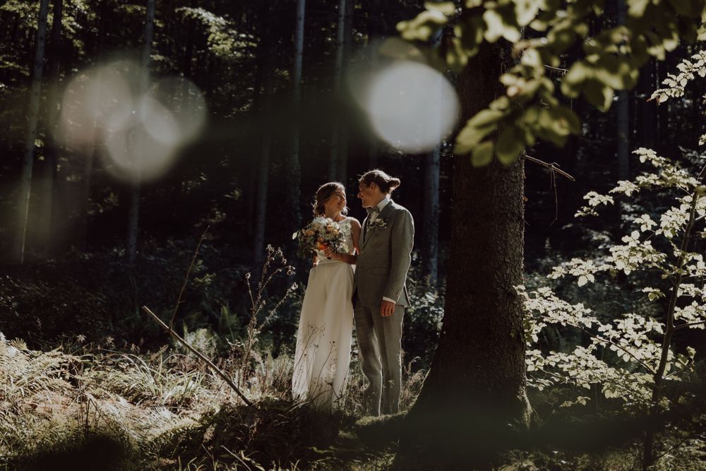 A couple stands together in dappled sunlight during their outdoor forest wedding ceremony.