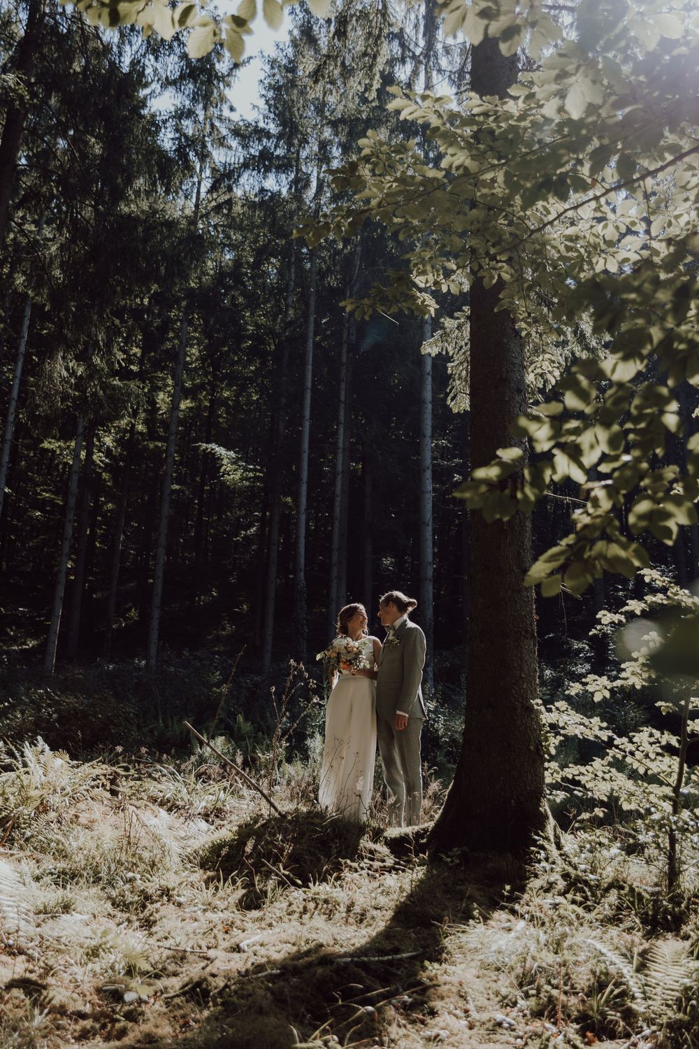 A couple in wedding attire standing together in a sunlit forest clearing.
